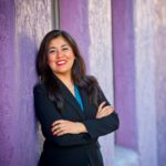 A woman standing in front of purple wall with her arms crossed.
